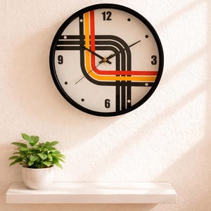 Könnte beinhalten: Eine runde Wanduhr mit schwarzem Rahmen und weißem Zifferblatt. Die Uhr hat ein Retro-Design mit schwarzen, roten und orangefarbenen Streifen. Sie ist an einer weißen Wand über einem weißen Regal mit einer Topfpflanze angebracht.