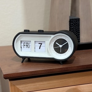 May include: A black and white retro flip clock displaying "FRI 7". The clock has a round analog clock face on the right side. The clock is on a wooden surface.
