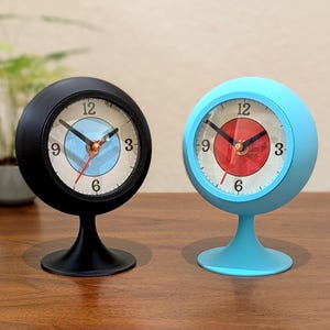 May include: Two retro-style desk clocks. One is black, the other is light blue. Both have a round face with a white background, black numbers, and red and blue hands. The clocks sit on a pedestal base.