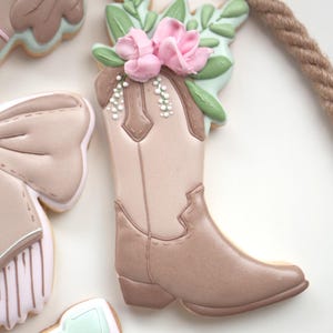 Può includere: Una collezione di biscotti decorati a tema western. Il punto focale è un biscotto a forma di stivale da cowboy adornato con fiori rosa e foglie verdi. Altri biscotti includono un ferro di cavallo e un fiocco.