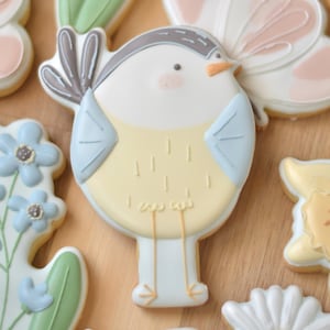 May include: A collection of decorated sugar cookies in various shapes. The central cookie is a bird with a yellow body, blue wings, and a gray head. Other cookies include flowers and a butterfly, all with pastel colors.