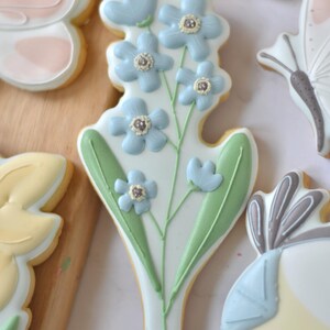 Può includere: Un biscotto di zucchero decorato a forma di bouquet di nontiscordardimé blu con foglie verdi. Il biscotto è bianco con fiori azzurri e un bordo giallo. Altri biscotti decorati sono visibili sullo sfondo.