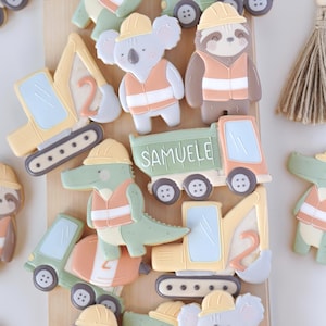 Può includere: Assortimento di biscotti a tema costruzione, con personaggi di animali come bradipi, koala e alligatori, tutti con giubbotti di sicurezza e caschi. I veicoli da costruzione includono un autocarro con cassone ribaltabile con il nome "Samuele" e un escavatore. I biscotti sono nei toni del giallo, arancione, verde e marrone.