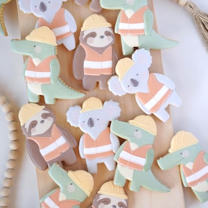 Può includere: Una collezione di biscotti decorati con personaggi di animali come alligatori, bradipi e koala. Ogni biscotto indossa un giubbotto e un casco da operaio. I biscotti sono nei toni del bianco, marrone, verde e arancione.