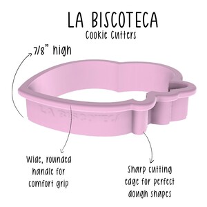May include: A light pink cookie cutter in the shape of a heart with a wide, rounded handle for a comfortable grip. The cutter is 7/8" high and has a sharp cutting edge for perfect dough shapes. The text "LA BISCOTECA Cookie Cutters" is visible.
