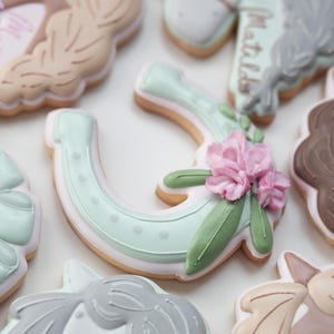 May include: Close-up of decorated sugar cookies with a horse theme. The cookies are in various shapes, including a horseshoe decorated with pink flowers and green leaves. Other cookies feature horse heads and manes, with pastel colors.