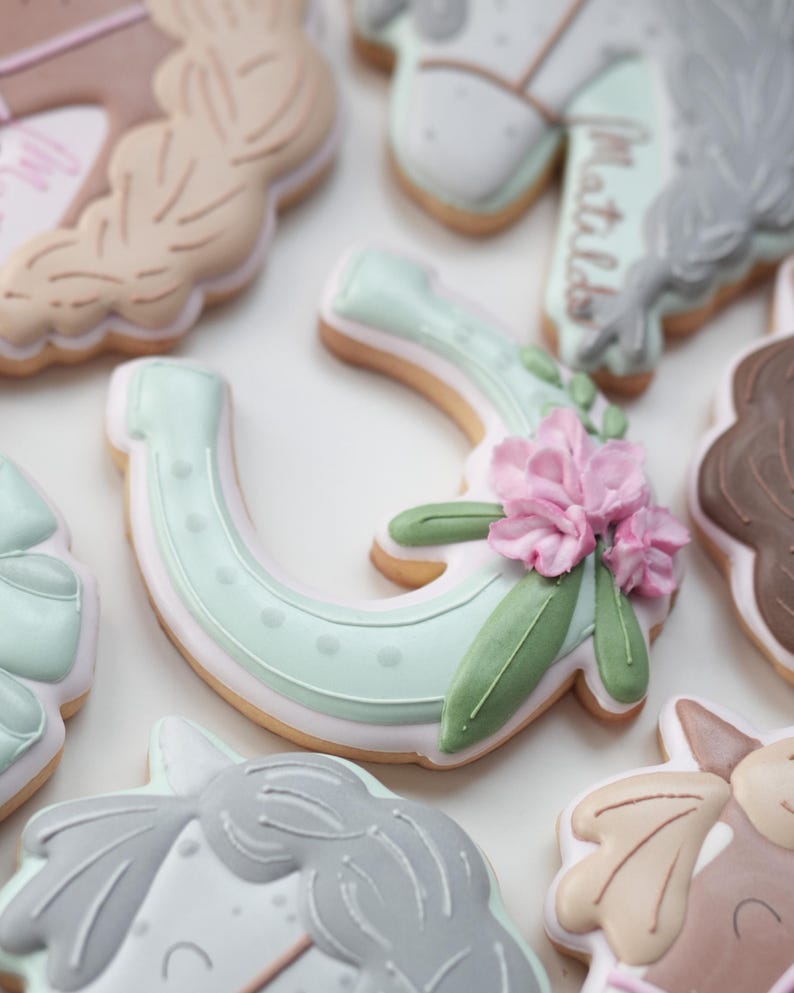 May include: Close-up of decorated sugar cookies with a horse theme. The cookies are in various shapes, including a horseshoe decorated with pink flowers and green leaves. Other cookies feature horse heads and manes, with pastel colors.