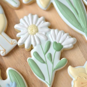 Può includere: Primo piano di biscotti di zucchero decorati a forma di fiori. I biscotti presentano margherite bianche con centri gialli, foglie e steli verdi. Altri biscotti nell'immagine includono narcisi e un piccolo fiore blu. I biscotti sono su una superficie di legno marrone chiaro.