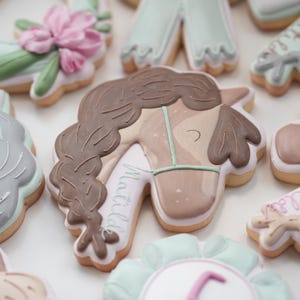 May include: Assortment of decorated sugar cookies. The cookies feature designs such as a horse head with a braided mane, a floral arrangement, and a number 5. The cookies are decorated with pastel colors and intricate details.