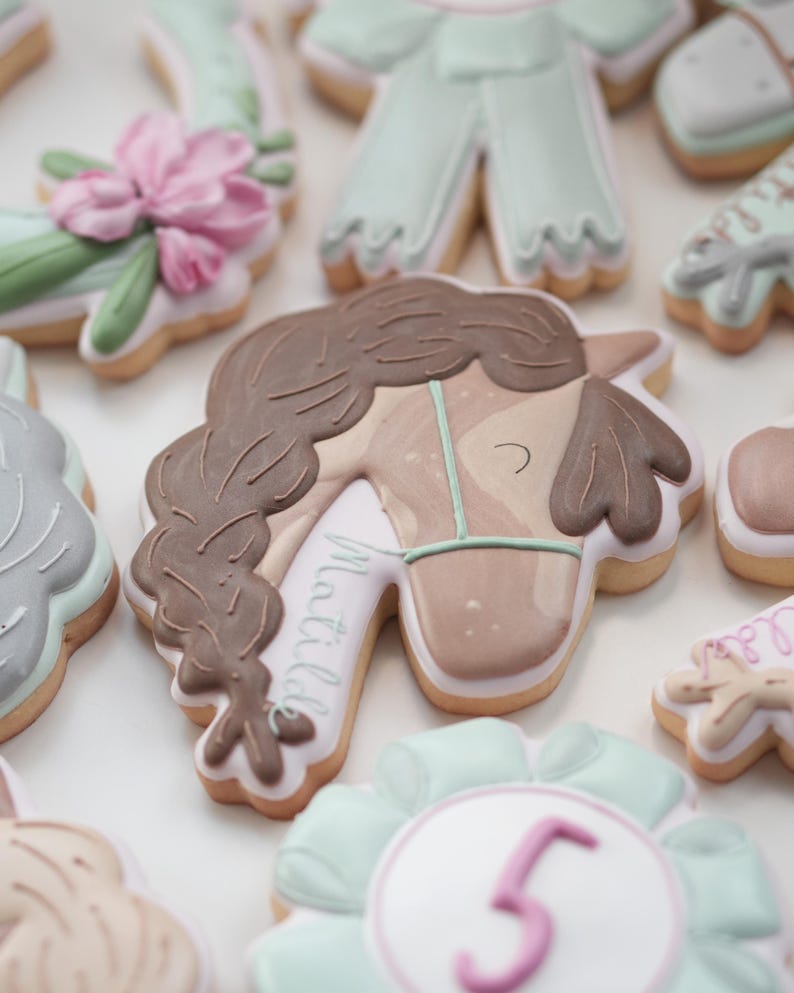 May include: Assortment of decorated sugar cookies. The cookies feature designs such as a horse head with a braided mane, a floral arrangement, and a number 5. The cookies are decorated with pastel colors and intricate details.
