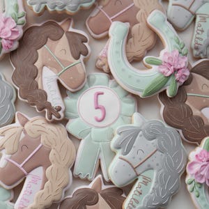 May include: Assortment of horse-themed cookies, including horse heads with braided manes, horseshoes, and a ribbon with the number 5. The cookies are decorated with pastel colors, including pink, brown, and mint green, and the name "Matilda" is written on some.