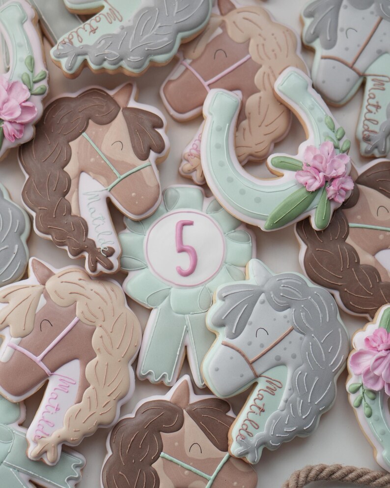 May include: Assortment of horse-themed cookies, including horse heads with braided manes, horseshoes, and a ribbon with the number 5. The cookies are decorated with pastel colors, including pink, brown, and mint green, and the name "Matilda" is written on some.