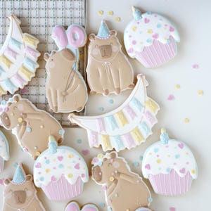 Può includere: Una collezione di biscotti decorati raffiguranti capibara color marrone chiaro, palloncini rosa a forma di dieci, cupcake color pastello con candeline e uno striscione a mezzaluna con il testo HAPPY BIRTHDAY in azzurro, giallo e rosa pastello.