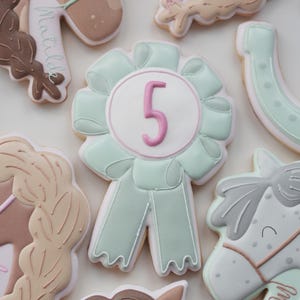 May include: Assortment of horse-themed cookies, including a large, light green ribbon cookie with a pink number 5. Other cookies feature horse heads, braids, and a horseshoe, all decorated with pastel colors and intricate details. The cookies are likely for a birthday.
