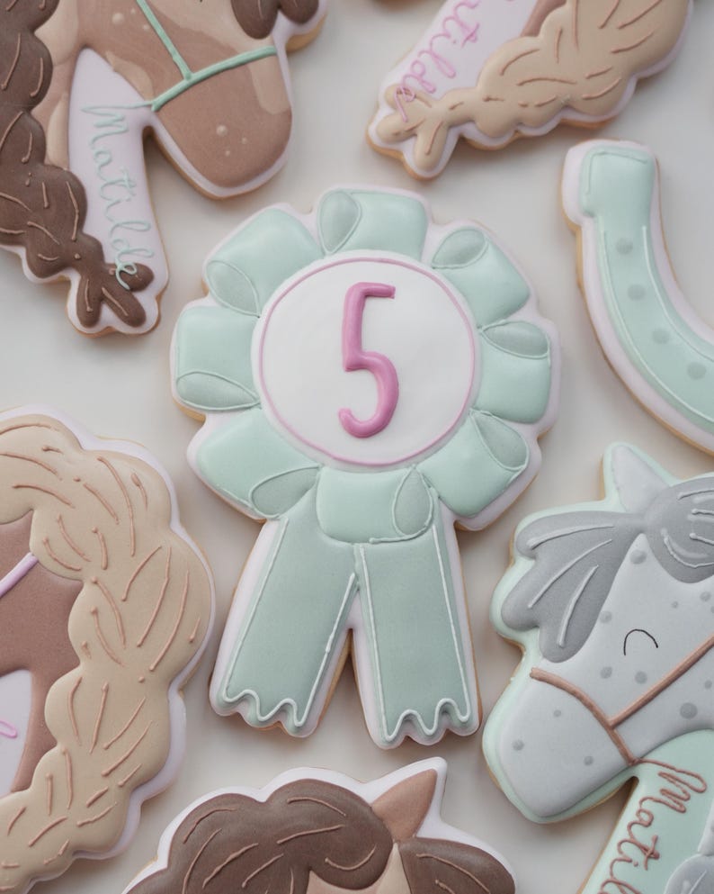May include: Assortment of horse-themed cookies, including a large, light green ribbon cookie with a pink number 5. Other cookies feature horse heads, braids, and a horseshoe, all decorated with pastel colors and intricate details. The cookies are likely for a birthday.
