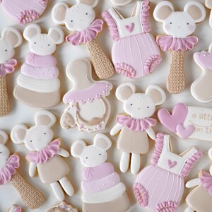 May include: An assortment of decorated sugar cookies in a baby shower theme. The cookies include mouse faces, baby rattles, baby rompers, and hearts. The cookies are decorated with white, pink, and tan icing.