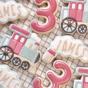 May include: Decorated sugar biscuits in the shape of a train, the number 3, and clouds with the name "JAMES" in white icing. The biscuits are decorated with pink, white, and light blue icing. The biscuits are arranged on a metal cooling rack.