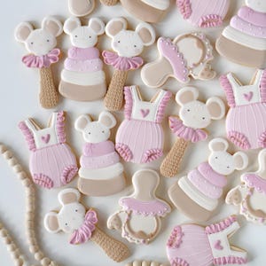 Op de afbeelding: Een assortiment baby-thema koekjes, waaronder muisvormige rammelaars, babyrompers met hartdetails en fopspeenkoekjes. De koekjes zijn versierd met roze, witte en beige glazuur.