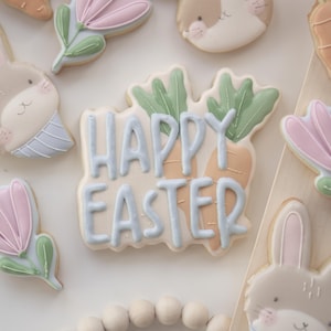 Könnte beinhalten: Ostergebäck mit pastellfarbenem Zuckerguss. Der zentrale Keks zeigt "Happy Easter" in hellblauem Zuckerguss, verziert mit Karotten- und Blattdetails. Weitere Kekse zeigen Hasen- und Blumenformen, alle in zarten Pastellfarben.