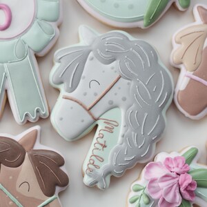 May include: Assortment of decorated sugar cookies in a horse theme. The cookies feature a horse head with the name "Matilda", a ribbon with the number "5", and horseshoe shapes with floral accents. The cookies are decorated with white, gray, pink, and mint green icing.