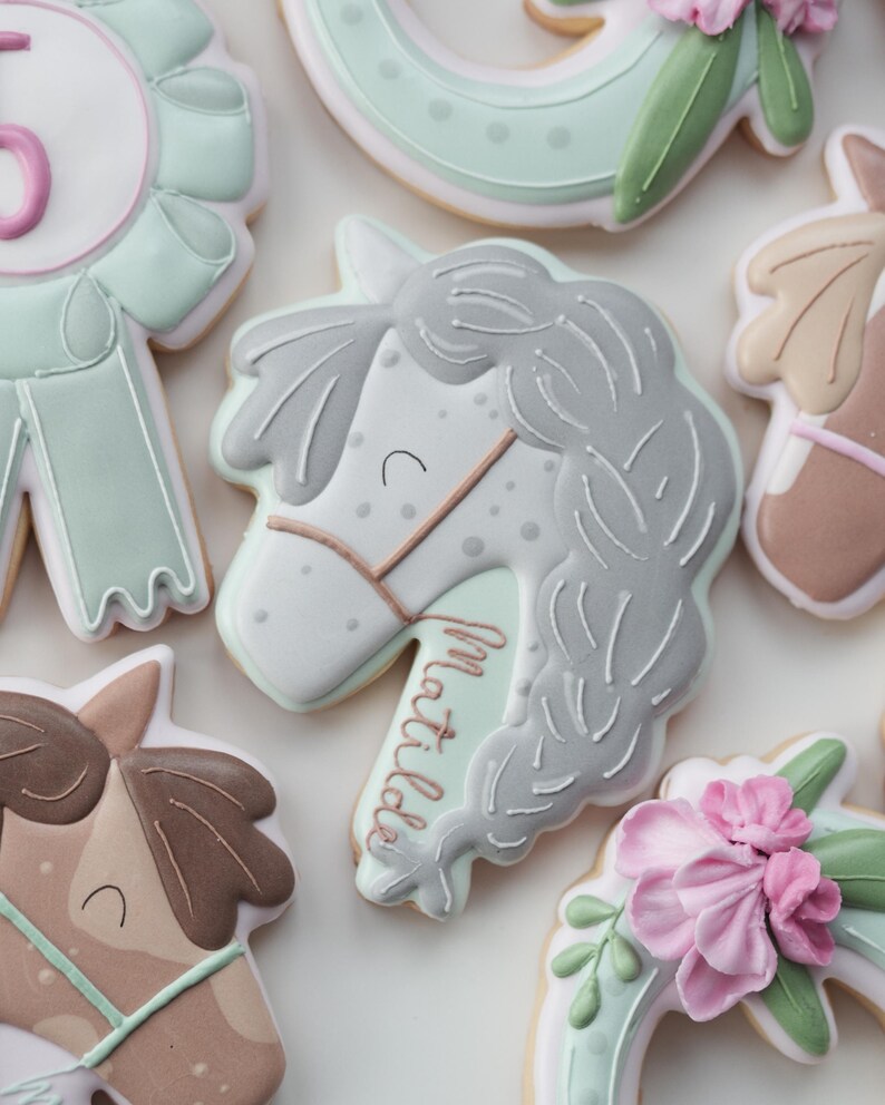 May include: Assortment of decorated sugar cookies in a horse theme. The cookies feature a horse head with the name "Matilda", a ribbon with the number "5", and horseshoe shapes with floral accents. The cookies are decorated with white, gray, pink, and mint green icing.