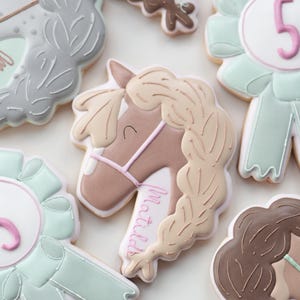 May include: Assortment of horse-themed cookies, including a horse head with a braided mane and the name "Matilda" in pink script. Other cookies are shaped like ribbons with the number 5, and rosettes in shades of gray, brown, and mint green.
