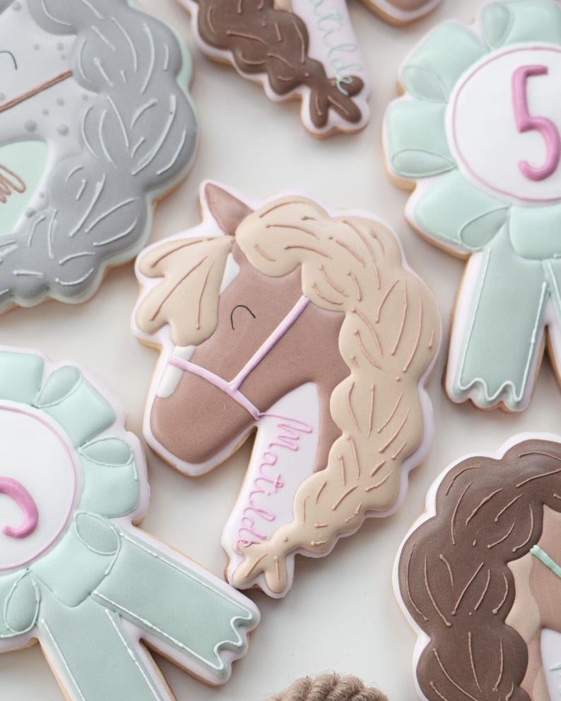 May include: Assortment of horse-themed cookies, including a horse head with a braided mane and the name "Matilda" in pink script. Other cookies are shaped like ribbons with the number 5, and rosettes in shades of gray, brown, and mint green.