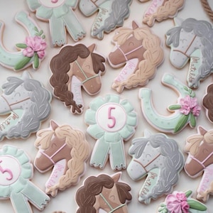 May include: An assortment of horse-themed cookies, including horse heads, horseshoes, and ribbons. The cookies feature pastel colors, with details like pink flowers and the number 5. Some cookies have the name "Matilda" written on them.