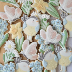 May include: A collection of decorated sugar cookies in the shapes of butterflies, birds, daisies, daffodils, and forget-me-nots. The cookies are primarily white, pastel pink, yellow, and blue, with green leaves. The cookies are arranged on a light brown surface.