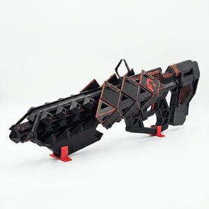 Könnte beinhalten: Ein schwarz-rotes Science-Fiction-Gewehr-Requisit mit geometrischem, kantigem Design, strukturiertem Lauf, markantem Visier und Schulterstütze. Die Waffe wird von zwei kleinen roten Ständern gehalten und zeigt eine detaillierte, facettierte Außenkonstruktion.
