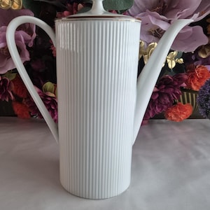 Op de afbeelding: Een witte keramische koffiepot met een geribbeld ontwerp. De pot heeft een lange, gebogen tuit en een handvat. Het deksel is bekroond met een kleine, ronde knop. De rand van de pot en het deksel zijn geaccentueerd met een gouden band. De achtergrond heeft kleurrijke bloemen.