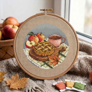 Puede incluir: Un bastidor de bordado circular de madera que contiene un diseño de punto de cruz de un pastel con rejilla, una taza rosa y manzanas rojas sobre una tabla de madera. La tela es de color tostado claro e incluye una hoja de otoño naranja.
