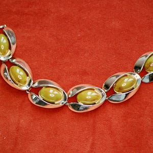 May include: A vintage bracelet featuring oval olive-green stones set in silver-tone metal. The bracelet has a series of linked, stylized oval shapes, creating a bold and elegant design. The clasp is a simple hook and eye closure. The bracelet is displayed on a red background.