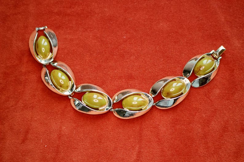 May include: A vintage bracelet featuring oval olive-green stones set in silver-tone metal. The bracelet has a series of linked, stylized oval shapes, creating a bold and elegant design. The clasp is a simple hook and eye closure. The bracelet is displayed on a red background.