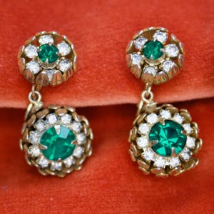 Puede incluir: Un par de pendientes colgantes vintage. Cada pendiente presenta una piedra central verde esmeralda rodeada de cristales claros y brillantes, engastada en metal dorado. Los pendientes tienen un dise&ntilde;o floral.