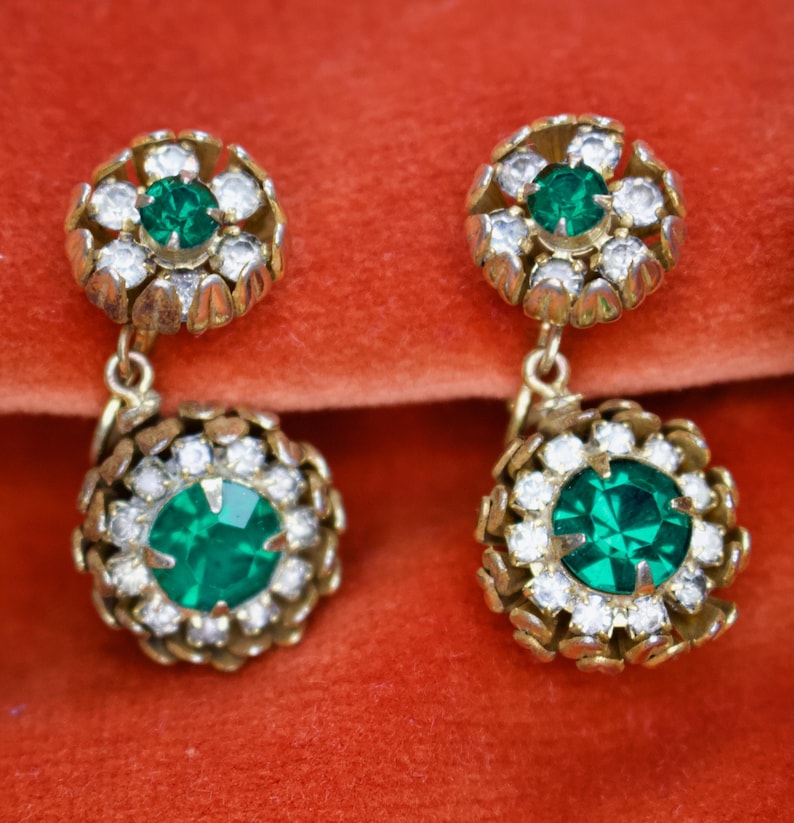 Puede incluir: Un par de pendientes colgantes vintage. Cada pendiente presenta una piedra central verde esmeralda rodeada de cristales claros y brillantes, engastada en metal dorado. Los pendientes tienen un dise&ntilde;o floral.