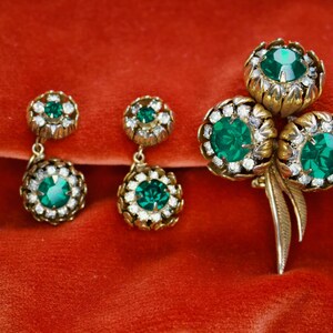 Puede incluir: Un conjunto de joyas vintage, que incluye un broche y dos pares de pendientes, con metal dorado y piedras preciosas de color verde esmeralda. El broche tiene tres piedras grandes, mientras que los pendientes tienen piedras m&aacute;s peque&ntilde;as y colgantes. Sobre un fondo rojo.