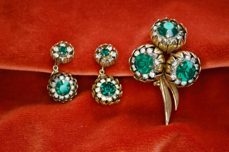 Puede incluir: Un conjunto de joyas vintage, que incluye un broche y dos pares de pendientes, con metal dorado y piedras preciosas de color verde esmeralda. El broche tiene tres piedras grandes, mientras que los pendientes tienen piedras m&aacute;s peque&ntilde;as y colgantes. Sobre un fondo rojo.
