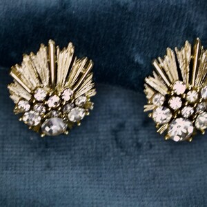 May include: A pair of gold-toned stud earrings. Each earring features a central cluster of clear, round rhinestones surrounded by smaller stones and radiating gold bars, set against a teal background.