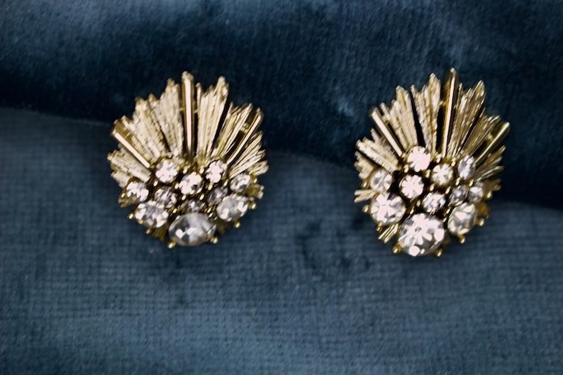 May include: A pair of gold-toned stud earrings. Each earring features a central cluster of clear, round rhinestones surrounded by smaller stones and radiating gold bars, set against a teal background.
