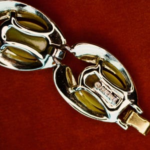 May include: Close-up of a gold-toned bracelet with oval-shaped links. The links have a textured design with a light green interior. The clasp is gold and the bracelet is on a red background. The bracelet is marked with the brand name "Ciner".