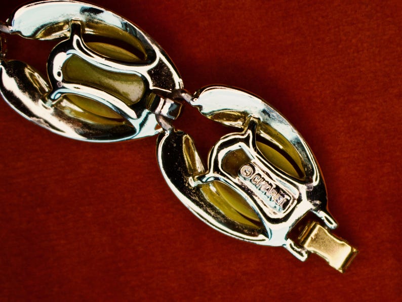 May include: Close-up of a gold-toned bracelet with oval-shaped links. The links have a textured design with a light green interior. The clasp is gold and the bracelet is on a red background. The bracelet is marked with the brand name "Ciner".