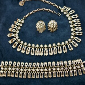 May include: A gold-toned jewelry set featuring a necklace, earrings, and bracelet. The necklace and bracelet have a repeating pattern of diamond-like accents. The earrings are round with a starburst design. The set is displayed on a dark blue surface.