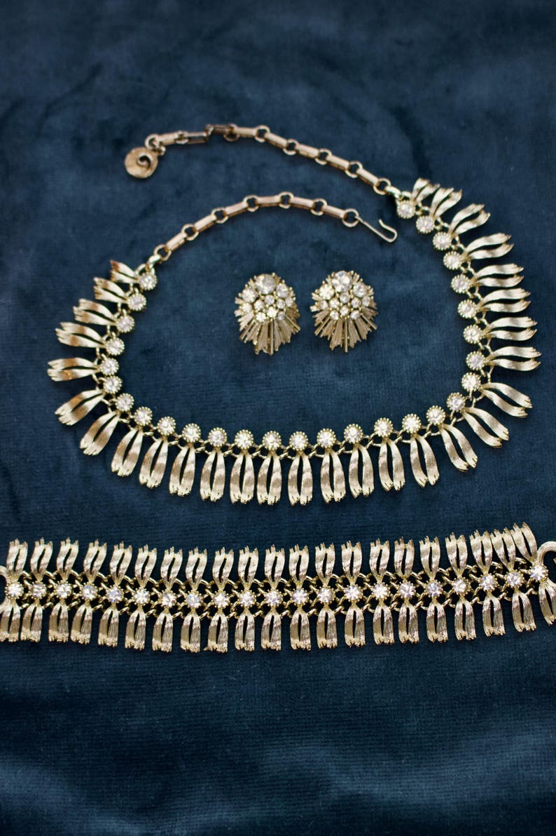 May include: A gold-toned jewelry set featuring a necklace, earrings, and bracelet. The necklace and bracelet have a repeating pattern of diamond-like accents. The earrings are round with a starburst design. The set is displayed on a dark blue surface.