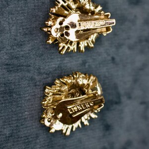 May include: Two gold-toned clip-on earrings with a textured, sunburst design. The earrings have a clip closure and the word "LISNERO" is stamped on the back. The earrings are set against a dark teal background.