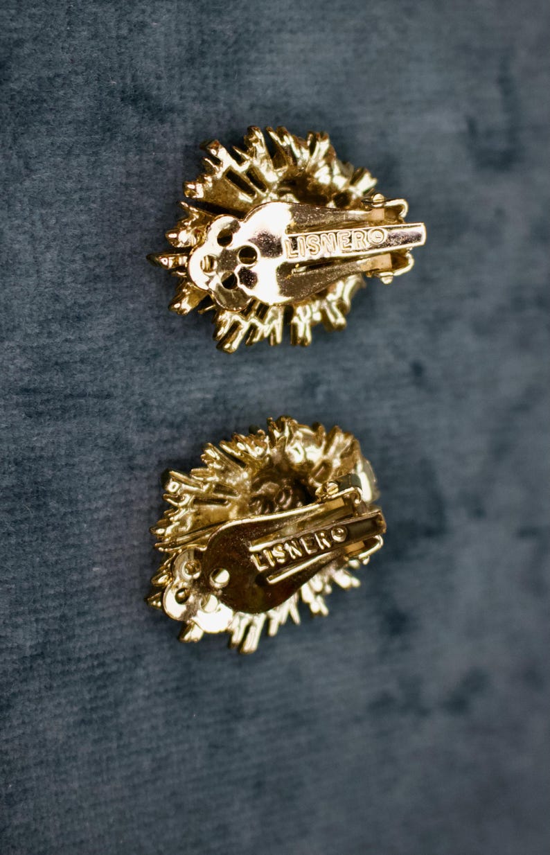 May include: Two gold-toned clip-on earrings with a textured, sunburst design. The earrings have a clip closure and the word "LISNERO" is stamped on the back. The earrings are set against a dark teal background.