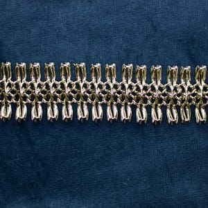 May include: A gold-toned bracelet with a repeating pattern of linked, stylized floral or teardrop shapes. The bracelet has a secure clasp and is displayed against a dark blue background. The jewelry piece is a classic accessory.