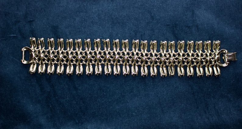 May include: A gold-toned bracelet with a repeating pattern of linked, stylized floral or teardrop shapes. The bracelet has a secure clasp and is displayed against a dark blue background. The jewelry piece is a classic accessory.