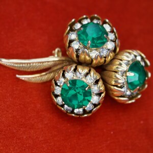 Puede incluir: Broche vintage dorado con tres racimos en forma de flor, cada uno con una piedra central verde esmeralda y cristales transparentes. El broche tiene un dise&ntilde;o de hojas y est&aacute; sobre un fondo rojo.