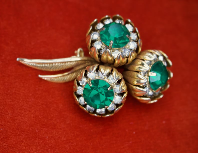 Puede incluir: Broche vintage dorado con tres racimos en forma de flor, cada uno con una piedra central verde esmeralda y cristales transparentes. El broche tiene un dise&ntilde;o de hojas y est&aacute; sobre un fondo rojo.